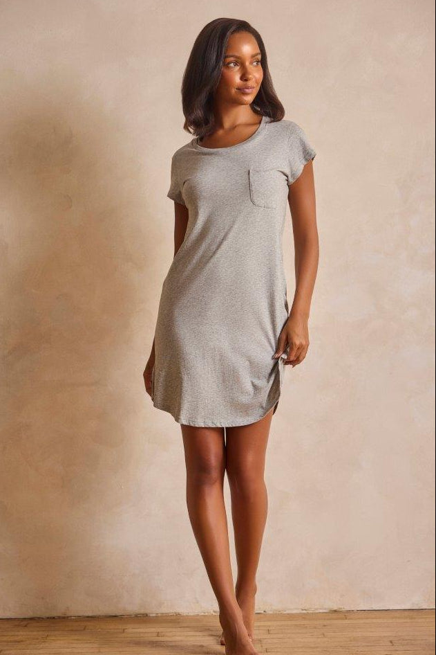 A woman modeling a light gray short-sleeve t-shirt dress with a chest pocket, standing against a textured beige wall.