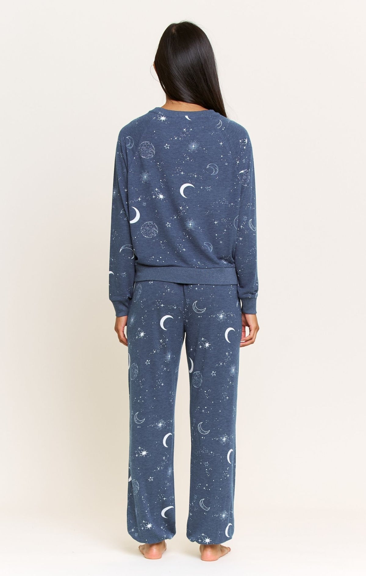 A woman viewed from the back wearing a matching navy blue pajama set with a white celestial print of crescent moons and stars, set against a plain off-white background.