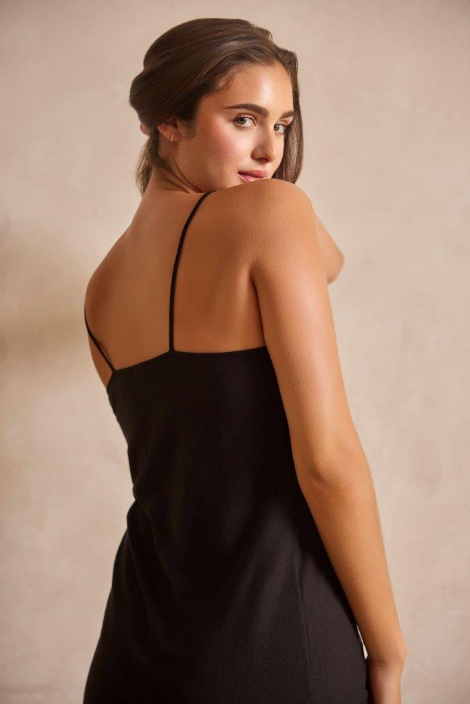A woman wearing a black spaghetti-strap slip dress, photographed from the back and side against a warm beige studio background.